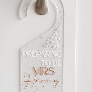 May include: Clear acrylic door hanger with silver hardware. The sign reads "PREPARING TO BE MRS Harvey PLEASE KNOCK" in white and copper-colored lettering. The hanger is decorated with glitter, pearls, and star-shaped accents.