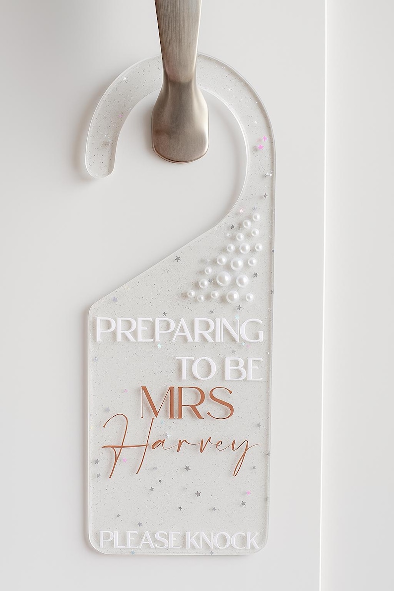 May include: Clear acrylic door hanger with silver hardware. The sign reads "PREPARING TO BE MRS Harvey PLEASE KNOCK" in white and copper-colored lettering. The hanger is decorated with glitter, pearls, and star-shaped accents.