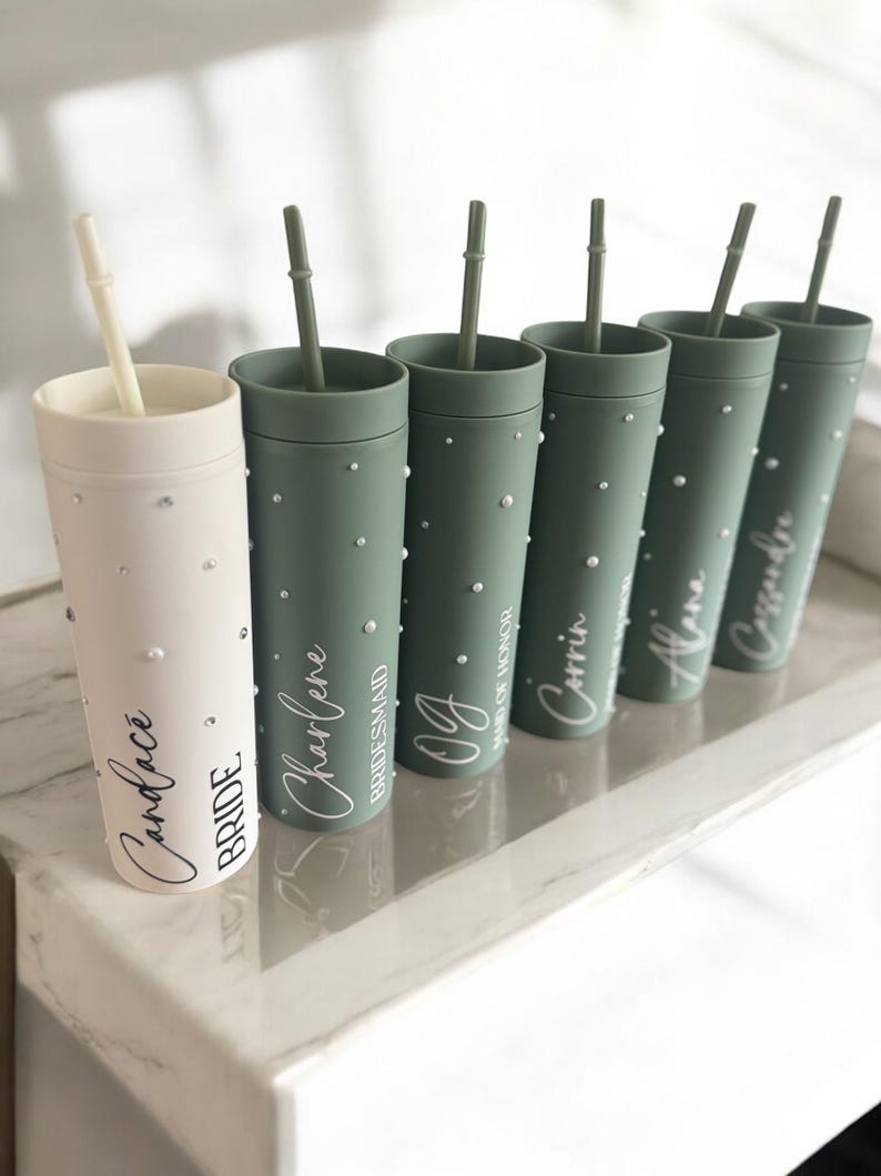 May include: Six green and white tumblers with straws and personalized names. The names are "Candace Bride", "Charlene Bridesmaid", "OG", "Maid of Honor", "Corrin", and "Alena".