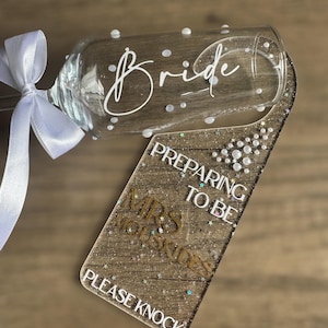 May include: A champagne flute with the word "Bride" in white script and white polka dots. A glittery door hanger reads "Preparing to be Mrs. [Last Name] Please Knock". A white ribbon is tied around the flute's stem.