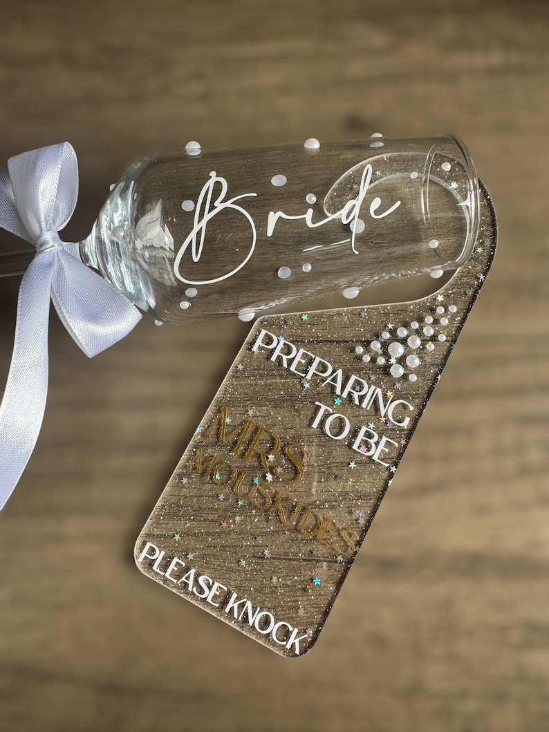 May include: A champagne flute with the word "Bride" in white script and white polka dots. A glittery door hanger reads "Preparing to be Mrs. [Last Name] Please Knock". A white ribbon is tied around the flute's stem.