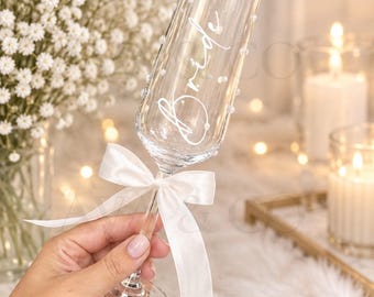 May include: A clear glass champagne flute featuring the word Bride in white cursive script, decorated with small white pearls adhered to the exterior surface, and accented with a white satin ribbon tied around the stem of the glass.