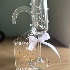 May include: A clear champagne flute with the word "Bride" in script. A clear acrylic door hanger with the text "Preparing to be Mrs. Mouskides, Please Knock" is attached with a white ribbon. The hanger has a glittery design.