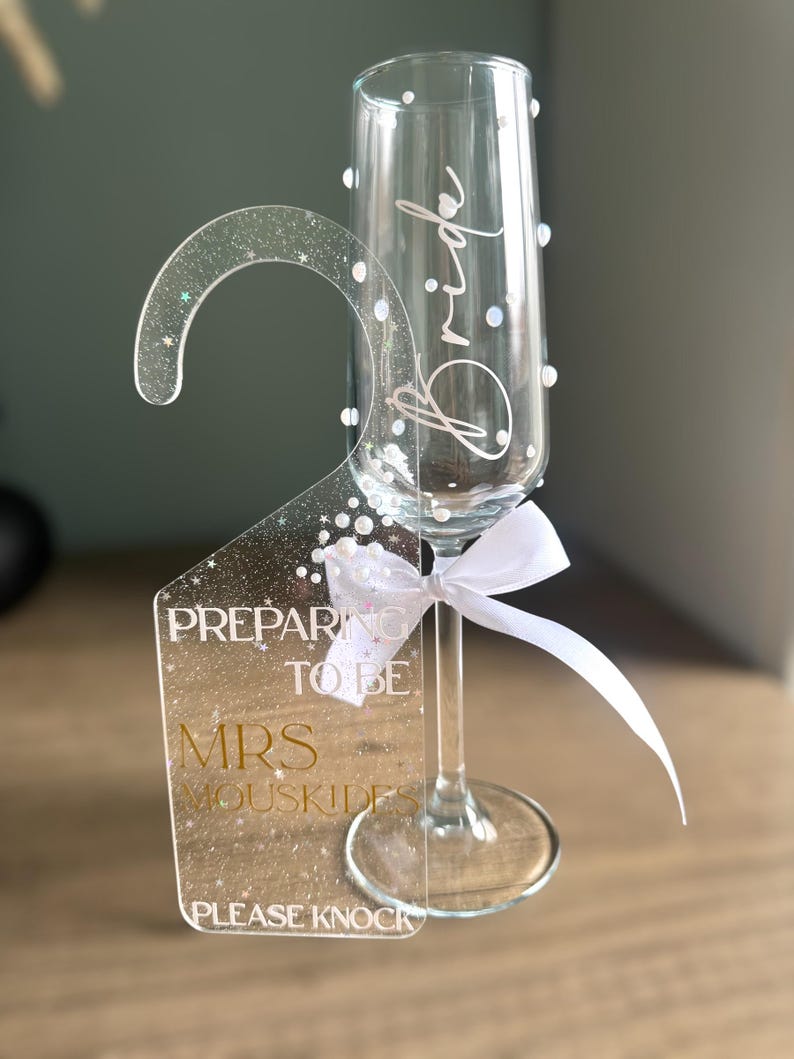 May include: A clear champagne flute with the word "Bride" in script. A clear acrylic door hanger with the text "Preparing to be Mrs. Mouskides, Please Knock" is attached with a white ribbon. The hanger has a glittery design.