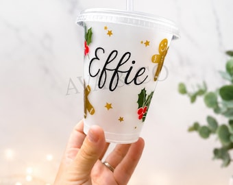 Puede incluir: Un vaso reutilizable de plástico transparente con tapa y pajita, que presenta el nombre Effie en letras cursivas negras. El vaso está decorado con hombres de jengibre, hojas de acebo verdes con bayas rojas y pequeñas estrellas doradas.