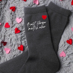 Könnte beinhalten: Ein Paar anthrazitfarbene Socken mit dem Text "Best Hinge match ever" in weißer Schrift und einem roten Herz. Rote und rosa herzförmige Papierstücke liegen auf einer grauen, strukturierten Oberfläche.
