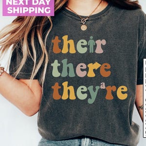 May include: A dark gray t-shirt with the words "their", "there", and "they're" printed in colorful, retro-style lettering.