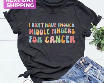 May include: A black short-sleeved t-shirt featuring the text I DON'T HAVE ENOUGH MIDDLE FINGERS FOR CANCER in a multi-coloured retro font. The shirt is paired with blue denim jeans and a pair of white low-top trainers with grey laces.