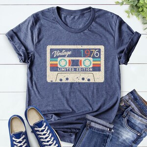 Op de afbeelding: Marineblauw t-shirt met een vintage cassettebandje ontwerp. De cassetteband grafiek toont de tekst "Vintage 1976" en "Limited Edition". De afbeelding toont ook blauwe sneakers en een spijkerbroek.