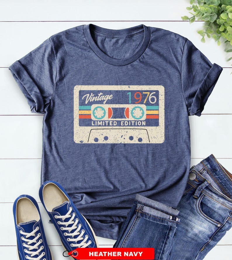Op de afbeelding: Marineblauw t-shirt met een vintage cassettebandje ontwerp. De cassetteband grafiek toont de tekst "Vintage 1976" en "Limited Edition". De afbeelding toont ook blauwe sneakers en een spijkerbroek.