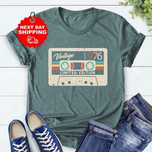 Op de afbeelding: Heather forest green t-shirt met een vintage cassettebandje ontwerp met de tekst "Vintage 1976 Limited Edition". De afbeelding toont ook een blauwe jeans en blauwe sneakers.