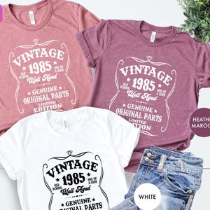 May include: Three t-shirts in heather maroon, white, and a dusty rose color, each with a vintage-style graphic. The graphic includes the text "VINTAGE 1985" and "Genuine Original Parts". Blue jeans are also visible.
