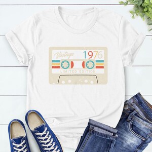 Op de afbeelding: Wit T-shirt met een vintage cassettebandje ontwerp met de tekst "Vintage 1976 Limited Edition". De afbeelding bevat retro kleuren. Blauwe sneakers en een spijkerbroek zijn ook zichtbaar.
