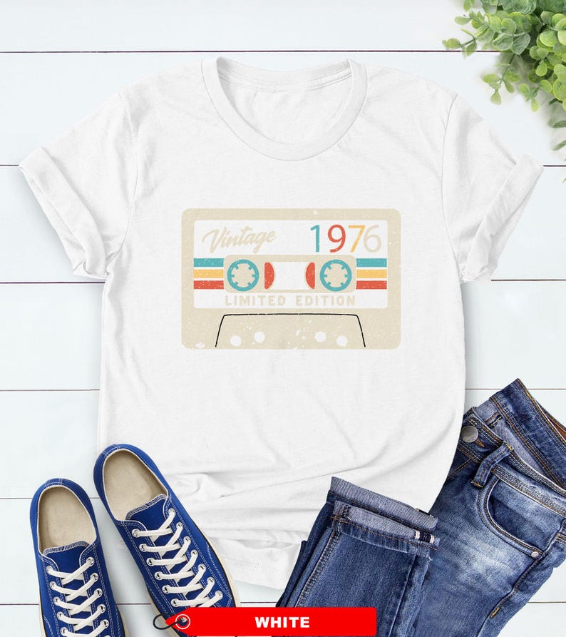 Op de afbeelding: Wit T-shirt met een vintage cassettebandje ontwerp met de tekst "Vintage 1976 Limited Edition". De afbeelding bevat retro kleuren. Blauwe sneakers en een spijkerbroek zijn ook zichtbaar.