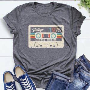 Op de afbeelding: Donkergrijze heather t-shirt met een vintage cassettebandje ontwerp met de tekst "Vintage 1976 Limited Edition". Het cassettebandje ontwerp heeft blauwe, rode en gele strepen. Het shirt wordt getoond met blauwe sneakers en een jeans.