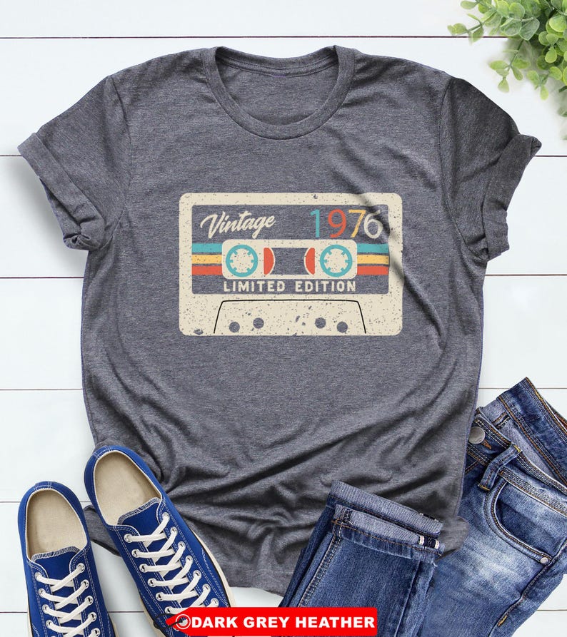 Op de afbeelding: Donkergrijze heather t-shirt met een vintage cassettebandje ontwerp met de tekst "Vintage 1976 Limited Edition". Het cassettebandje ontwerp heeft blauwe, rode en gele strepen. Het shirt wordt getoond met blauwe sneakers en een jeans.