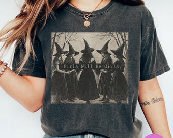 May include: Dark gray t-shirt featuring a sepia-toned graphic of five women in witch hats, with the text "Girls Will Be Girls." The design is centered on the shirt, creating a vintage aesthetic.