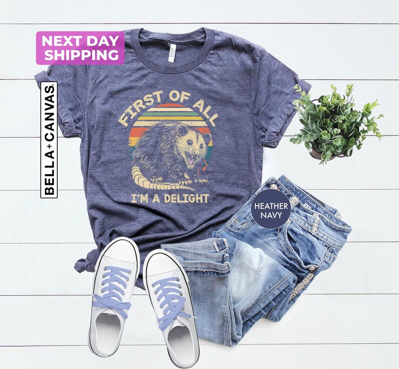May include: A heather navy blue t-shirt with a vintage-style graphic of a possum with the text "First of all, I'm a delight".