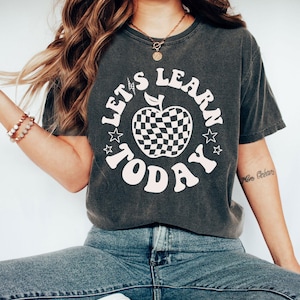 May include: A charcoal gray short-sleeve t-shirt featuring a white graphic of an apple with a checkered pattern. The text "LET'S LEARN TODAY" is printed in a curved white font around the apple, accompanied by small lightning bolt and star icons.