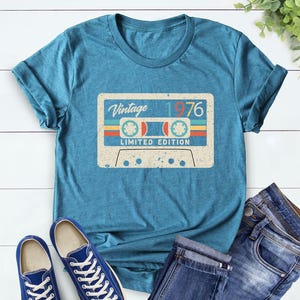 Op de afbeelding: Tealkleurig T-shirt met een vintage cassettebandje ontwerp. De afbeelding toont de tekst "Vintage 1976" en "Limited Edition". Blauwe sneakers en een jeans liggen naast het shirt op een witte houten achtergrond.
