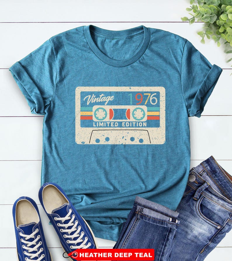 Op de afbeelding: Tealkleurig T-shirt met een vintage cassettebandje ontwerp. De afbeelding toont de tekst "Vintage 1976" en "Limited Edition". Blauwe sneakers en een jeans liggen naast het shirt op een witte houten achtergrond.