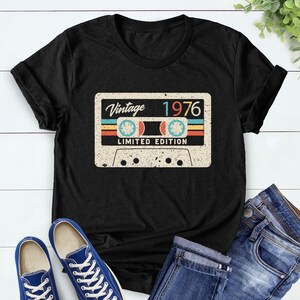Op de afbeelding: Zwart T-shirt met een vintage cassettebandje ontwerp. Het ontwerp bevat de tekst "Vintage 1976 Limited Edition" in retro kleuren. Het shirt wordt gecombineerd met een blauwe jeans en blauwe sneakers.