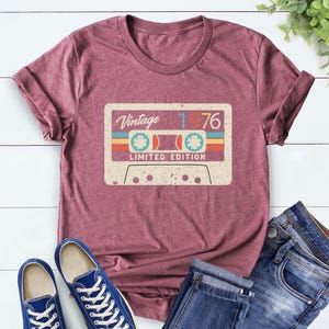 Op de afbeelding: Bordeauxrood t-shirt met een vintage cassettebandje ontwerp. De afbeelding bevat de tekst "Vintage 1976" en "Limited Edition" in retrostijl. Het t-shirt wordt gecombineerd met blauwe sneakers en een jeans.