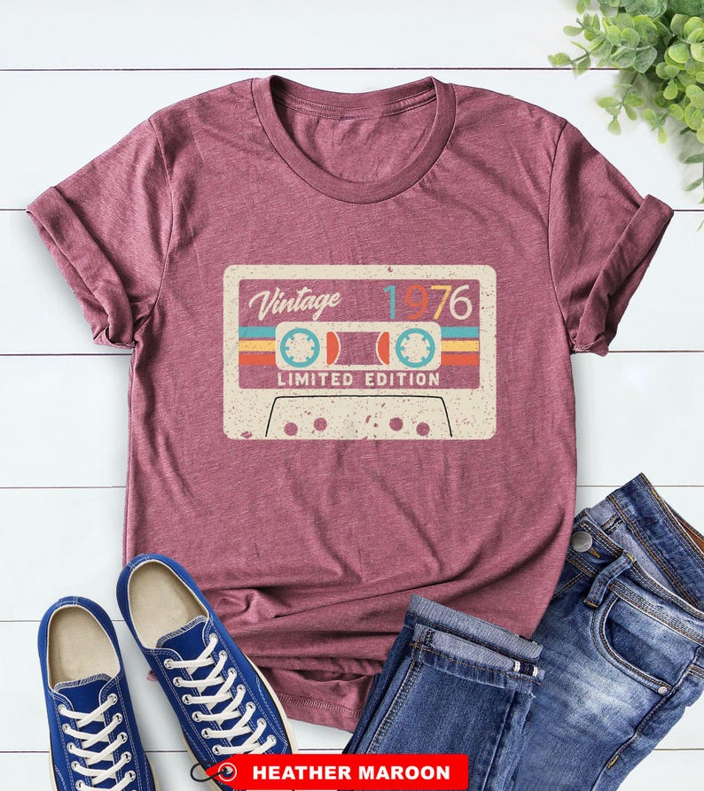Op de afbeelding: Bordeauxrood t-shirt met een vintage cassettebandje ontwerp. De afbeelding bevat de tekst "Vintage 1976" en "Limited Edition" in retrostijl. Het t-shirt wordt gecombineerd met blauwe sneakers en een jeans.