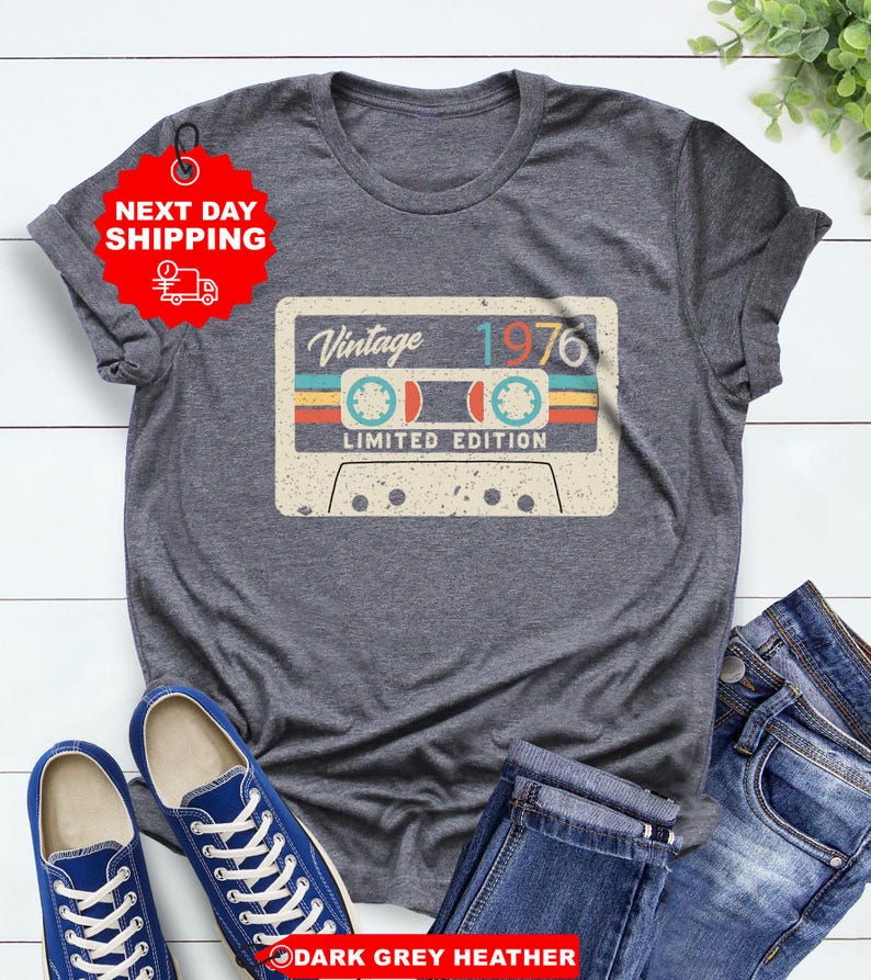 Op de afbeelding: Donkergrijze heather t-shirt met een vintage cassettebandje ontwerp en de tekst "Vintage 1976 Limited Edition". Het shirt wordt gecombineerd met een blauwe jeans en blauwe sneakers. Een rode sticker met "NEXT DAY SHIPPING" is bevestigd.
