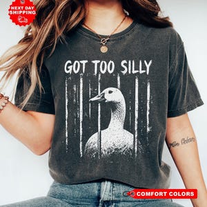 Może przedstawiać: Ciemnoszary t-shirt z krótkim rękawem z białym nadrukiem graficznym przedstawiającym gęś za pionowymi kratami. Napis GOT TOO SILLY jest nadrukowany na biało nad gęsią. Koszulka ma okrągły dekolt i wykończenie w stylu vintage z efektem sprania.