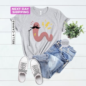 May include: A gray t-shirt with a cartoon worm wearing a mustache and holding a microphone. The worm is pink and has a lightning bolt behind it. The shirt is on a white wooden surface with a pair of white sneakers and blue jeans.