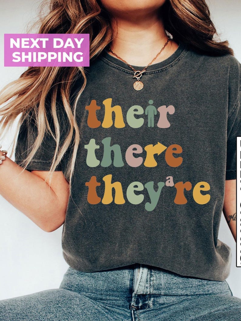 May include: A dark gray t-shirt with the words "their", "there", and "they're" printed in colorful, retro-style lettering.