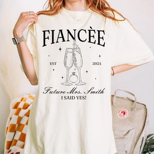Może przedstawiać: Kremowy t-shirt z czarnym napisem FIANCÉE, EST 2024, Future Mrs. Smith oraz I SAID YES! Grafika przedstawia dwa stykające się kieliszki do szampana oraz pierścionek zaręczynowy z diamentem umieszczony centralnie pod kieliszkami.