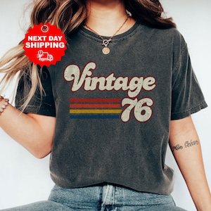 May include: Dark grey heather t-shirt featuring the text "Vintage 76" in a retro style. The design incorporates horizontal stripes in red, orange, yellow, and blue. The shirt has a crew neck and short sleeves.