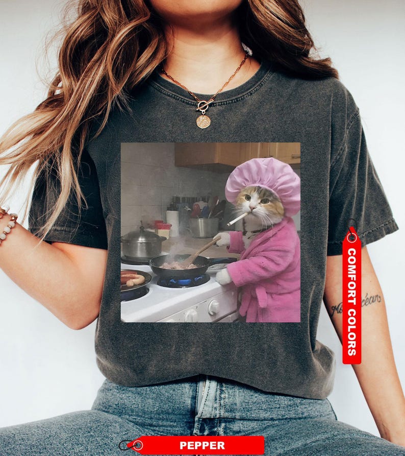 May include: Dark grey t-shirt featuring a humorous image of a cat in a pink robe and chef's hat, smoking a cigarette while cooking. The shirt has a Comfort Colors tag and a Pepper tag.