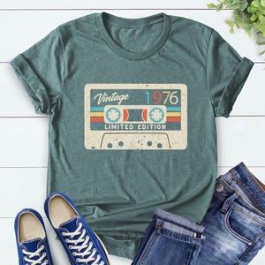 Op de afbeelding: Een tealkleurig T-shirt met een vintage cassettebandje ontwerp met de tekst "Vintage 1976 Limited Edition". Het shirt wordt gecombineerd met een blauwe jeans en blauwe sneakers. Het shirt is gemaakt van Heather Forest materiaal.