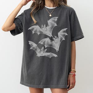 May include: Dark gray oversized t-shirt featuring a detailed white illustration of three bats in flight. The bats are depicted with intricate wing details, creating a vintage or gothic aesthetic. The shirt has a relaxed fit and a slightly faded appearance.