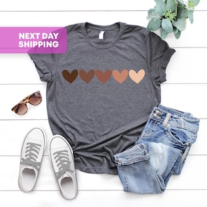 May include: A dark gray t-shirt with a row of five brown hearts of different shades across the chest. The shirt is laying flat on a white surface with a pair of white sneakers and blue jeans.