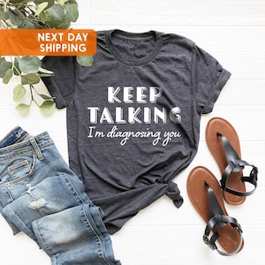 May include: Dark grey t-shirt with the text "KEEP TALKING I'm diagnosing you" in white. The shirt is styled with blue jeans and brown sandals. The image also includes a sprig of green leaves and an orange label.