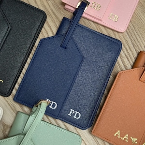 May include: A selection of personalised luggage tags in navy, black, pink, and orange. Each tag displays initials in white. The tags are made from a textured material. The tags are designed for travel.