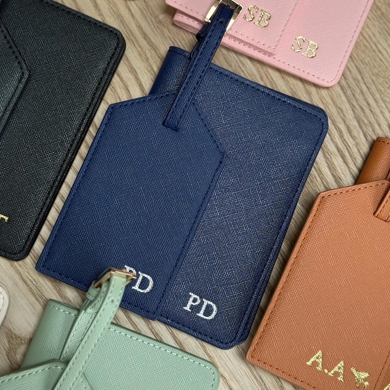 May include: A selection of personalised luggage tags in navy, black, pink, and orange. Each tag displays initials in white. The tags are made from a textured material. The tags are designed for travel.