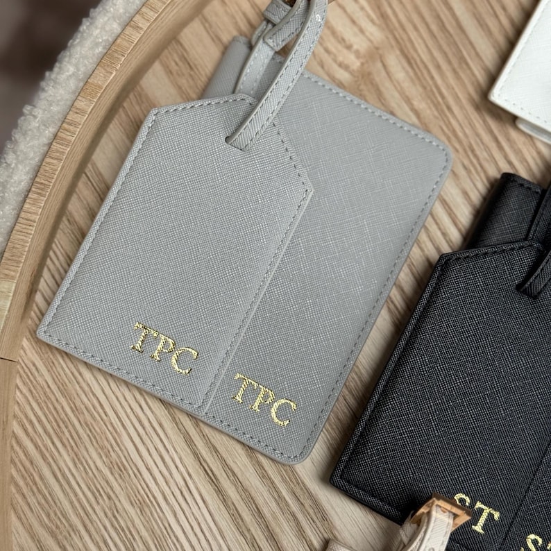 May include: Close-up of personalised luggage tags in grey and black. The grey tag has "TPC" in gold lettering. The black tag is partially visible. The tags are on a wooden surface.