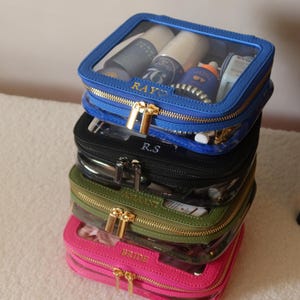 May include: Stack of five clear zippered cosmetic bags with different coloured exteriors. The bags are stacked from top to bottom: blue, black, green, pink. The top bag has the name "RAYO" embroidered in gold on the front. The second bag has the initials "R.S." embroidered in gold on the front. The third bag has the word "BEAUTY" embroidered in gold on the front. The fourth bag has the word "BRIDE" embroidered in gold on the front.