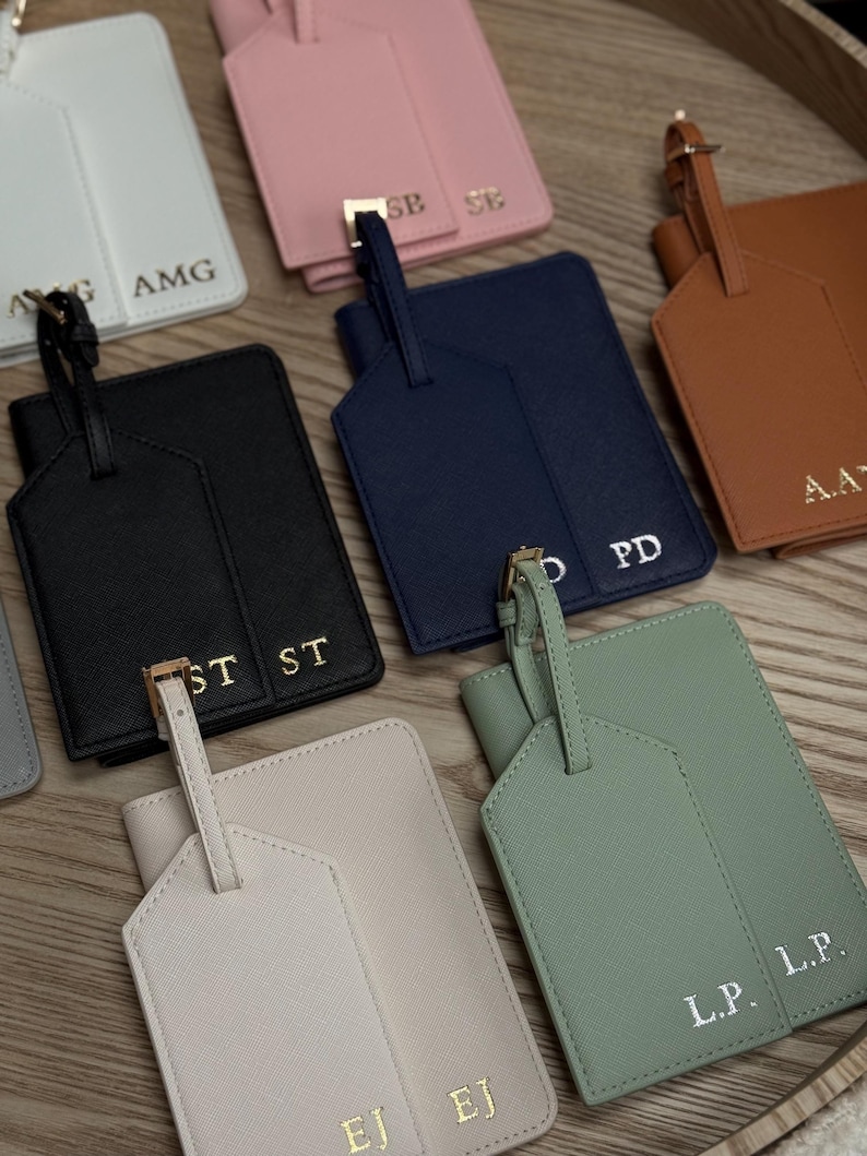 May include: A selection of personalised luggage tags and passport holders in colours such as black, pink, navy, and green. Each tag and holder has gold-coloured hardware and initials.