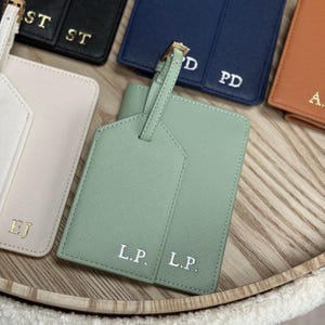 May include: A selection of personalised luggage tags and passport holders in colours such as black, navy, and mint green. Each item has initials in a contrasting colour, with a gold-coloured buckle on the luggage tag. The items are arranged on a wooden tray.