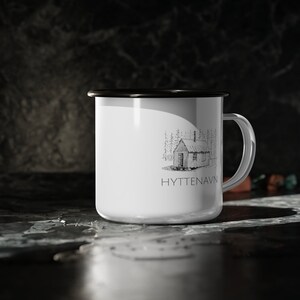 May include: White enamel mug with a black rim and handle. The mug features a black and white illustration of a cabin in the woods with the text "HYTTENAVN" below it.