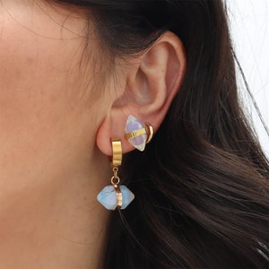 Peut inclure: Boucles d'oreilles créoles dorées avec un pendentif en cristal géométrique et irisé. Les boucles d'oreilles ont une bande dorée autour du cristal et un anneau doré. Le cristal est bleu clair et blanc.