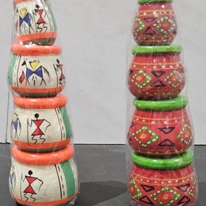 May include: Two stacks of hand-painted wooden pots. One stack features white pots with orange rims and green stripes, adorned with stick figure designs in red, blue, and yellow. The other stack has red pots with green rims and geometric patterns.