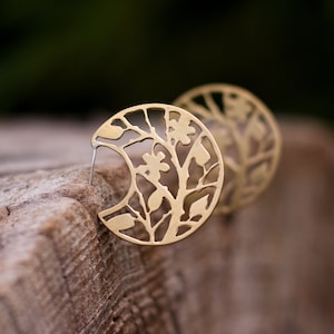 Round brass earrings with tree branches motif cut out are balancing on the round piece of wood. Earrings are shiny and golden, but not polished, they have a soft matte finish. Their pattern is tree branches with leaves and flowers.