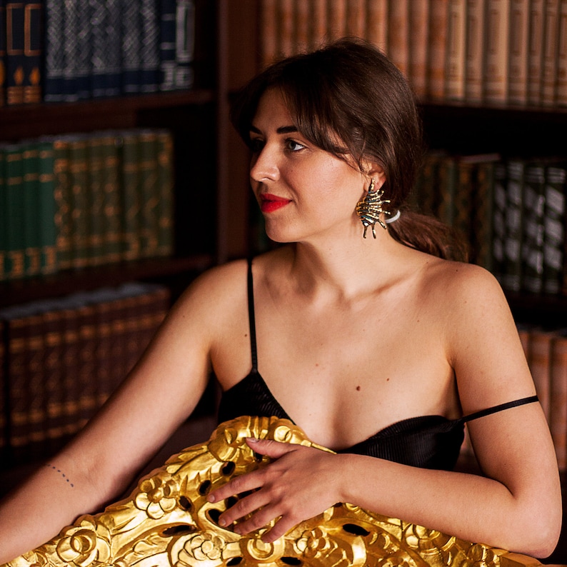 Young woman in bold golden metal earrings in the form of elongated spikey shells. Earrings have natural blue patina in the hollows of the metal. Shell studs are quite long, the hang along the neck without movement.